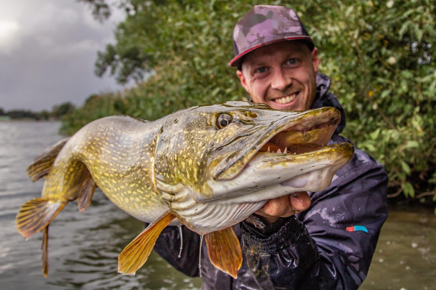 Pêcheur souriant tenant un gros brochet au bord d’une gravière en automne