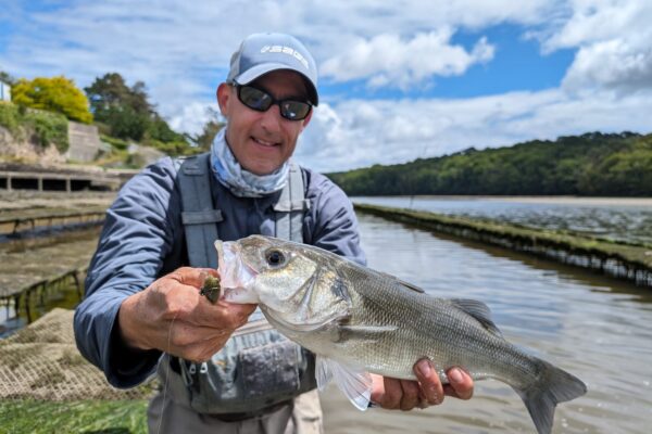 Jean-Baptiste Vidal tenant un bar pris à la mouche devant des parcs à huîtres, à marée basse en Bretagne