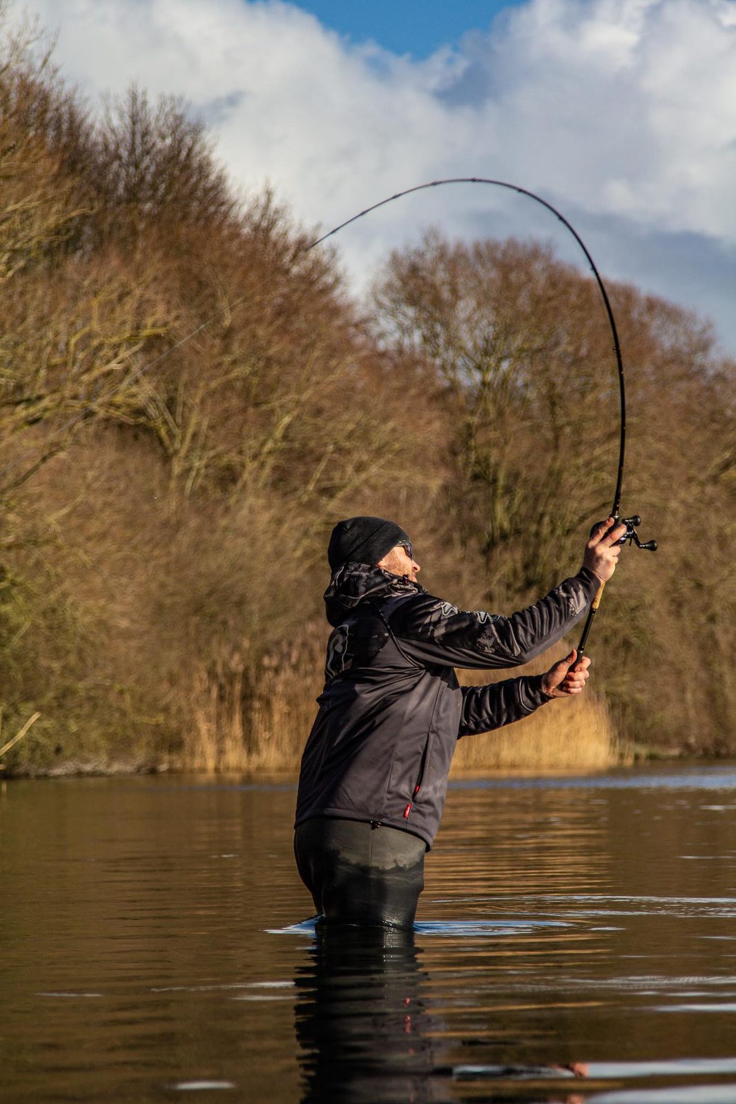 Pêcheur de brochet en wading dans une gravière, de l’eau jusqu’à la taille, canne pliée sur un poisson