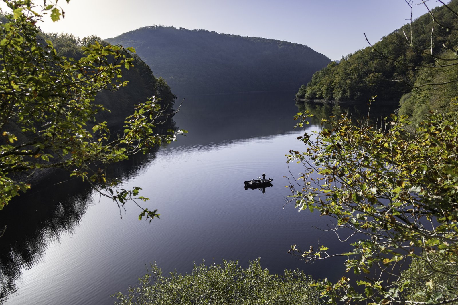 Pêcheur en barque sur un lac de barrage encaissé de Nouvelle-Aquitaine au petit matin