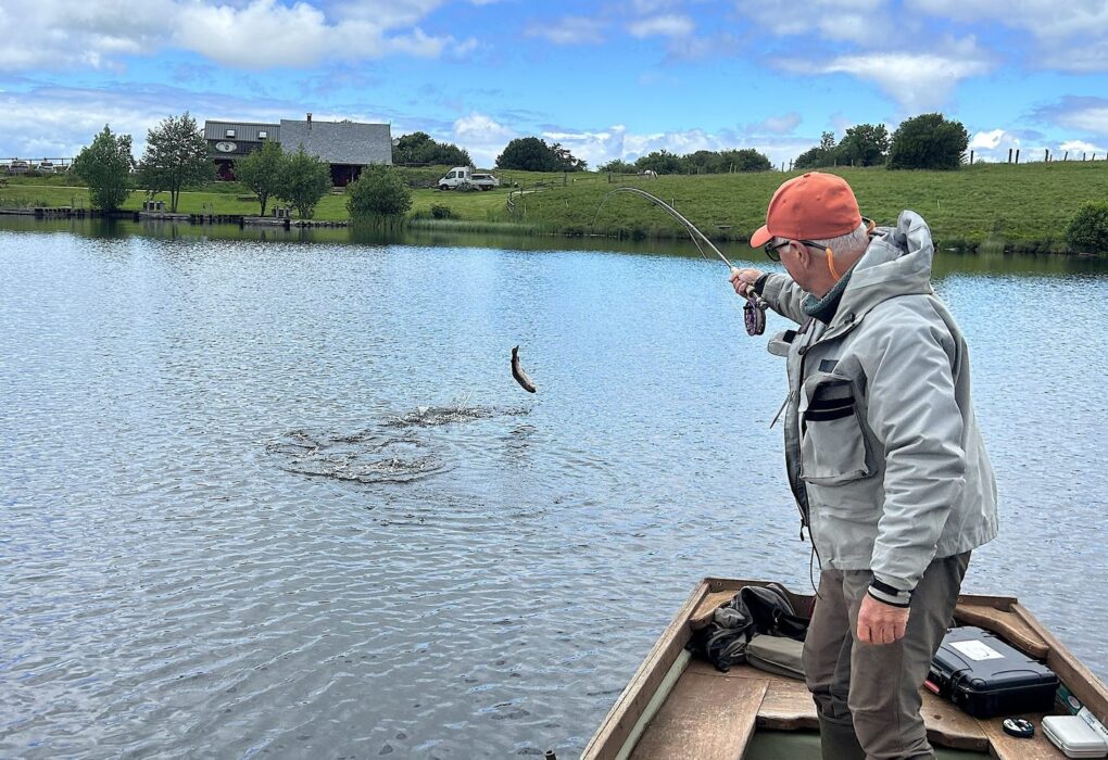 Pêcheur à la mouche en barque sur le lac de Veirières, une truite arc-en-ciel sautant hors de l’eau au bout de la ligne