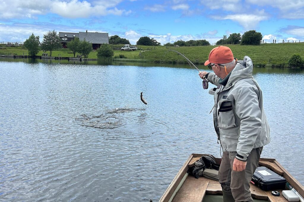 Pêcheur à la mouche en barque sur le lac de Veirières, une truite arc-en-ciel sautant hors de l’eau au bout de la ligne